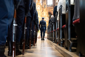 kolorowe zdjęcie policjanta stojącego pomiędzy rzędami ławek, wykonane za jego plecami