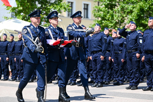 kolorowe zdjęcie maszerujących policjantów Kompani Honorowej z szablami i flagą RP, w tle policjanci oddają honory