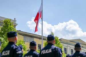 kolorowe zdjęcie przedstawiające stojących policjantów, w tle maszt z flagą RP
