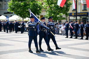 kolorowe zdjęcie przedstawiające policjantów Kompani Honorowej maszerujących ze sztandarem oraz szablami, w tle policjanci oddający honory