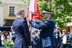 Kolorowe zdjęcie przedstawiające Generała Policji oraz mężczyzn w garniturze stojących przy sztandarze
