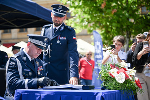 zdjęcie przedstawiające Generała Policji składającego podpis na dokumencie, w tle osoby fotografujące