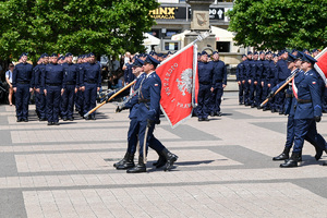 kolorowe zdjęcie policjantów maszerujących ze sztandarem, w tle policjanci stoją na baczność