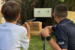 kolorowe zdjęcie przedstawiające chłopca strzelającego z wiatrówki do tarczy strzeleckiej znajdujący się w towarzystwie umundurowanego policjanta