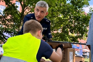 kolorowe zdjęcie przedstawiające chłopca strzelającego z wiatrówki do tarczy strzeleckiej znajdujący się w towarzystwie umundurowanego policjanta