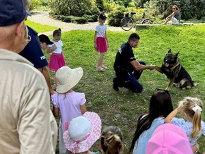 Kolorowe zdjęcie przedstawiające umundurowanego policjanta znajdującego się w towarzystwie owczarka niemieckiego, który podaje policjantowi łapę. W pobliżu znajdują się dzieci.
