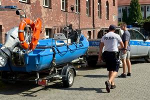 Kolorowe zdjęcie policjanta policji wodnej, w tle radiowóz z przyczepą, na której znajduje się łódź motorowa.