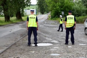 policjant i strażnik ochrony kolei stojący tyłem, przed nimi idąca policjantka, w tle widać przejazd kolejowy