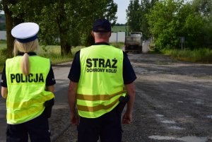 policjantka i pracownik straży ochrony kolei ustawieni tyłem od pasa w górę, w tle przejazd kolejowy