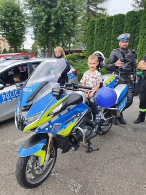 Chłopiec siedzący na motocyklu policyjnym, obok radiowóz oraz umundurowany policjant ruchu drogowego
