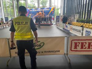 Policjant sw odblaskowej kamizelce podczas zabezpieczenia imprezy sportowej. W tle, w hali sportowej,  na podium zawodnicy