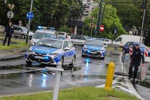 dwa policyjne radiowozy oraz samochód straży miejskiej z włączonymi sygnałami świtlnymi na jednej z krakowskich dróg. Po lewej stronie widać umundurowanego policjanta. W tle widać samochodowy ruch uliczny