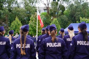 Na pierwszym planie nowoprzyjęci policjanci stojący tyłem do fotografującego w dwuszeregu. W tle flaga Polski powiewająca na maszcie.