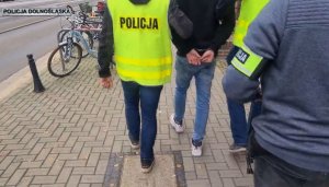 Zdjęcie przedstawia policjantów prowadzących zatrzymanego mężczyznę chodnikiem do radiowozu. Policjanci mają kamizelki odblaskowe i opaskę na ramieniu z napisem POLICJA. Zatrzymany ma ręce zapięte kajdankami z tyłu.