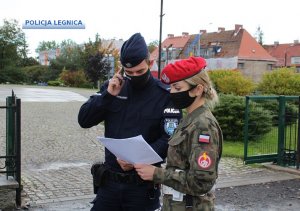 Na zdjęciu widoczny jest policjant w mundurze, który trzyma przy uchu telefon komórkowy. Obok niego stoi żołnierz.