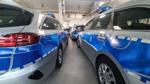 Na zdjęciu dwa nowe policyjne radiowozy stojące tyłem marki KIA, koloru srebrno-niebieskiego.