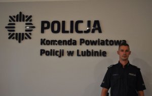 Na zdjęciu policjant Gaziński na tle napisu Komenda Powiatowa Policji w Lubinie.