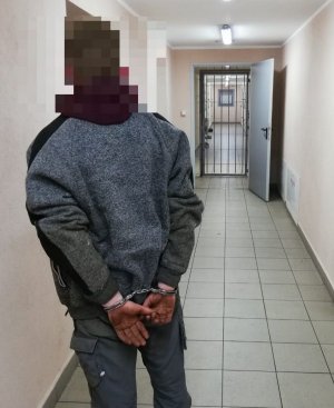 Na zdjęciu zatrzymany mężczyzna, skuty w kajdanki, stojący w holu budynku policji.