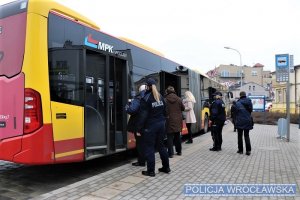 Zdjęcie przedstawia przystanek autobusowy i wsiadających do pojazdu pasażerów. Jest tam też policjantka w mundurze.