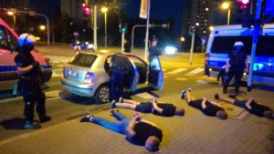 zdjęcie kolorowe: policjanci katowickiego oddziału prewencji nadzorujący 4 zatrzymanych pseudokibiców, którzy leżą na chodniku z założonymi kajdankami na ręce trzymane z tyłu