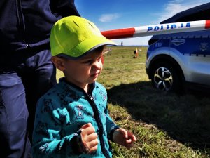 3-letni chłopiec w czapeczce z logo policyjnej kampanii, obok policyjny radiowóz