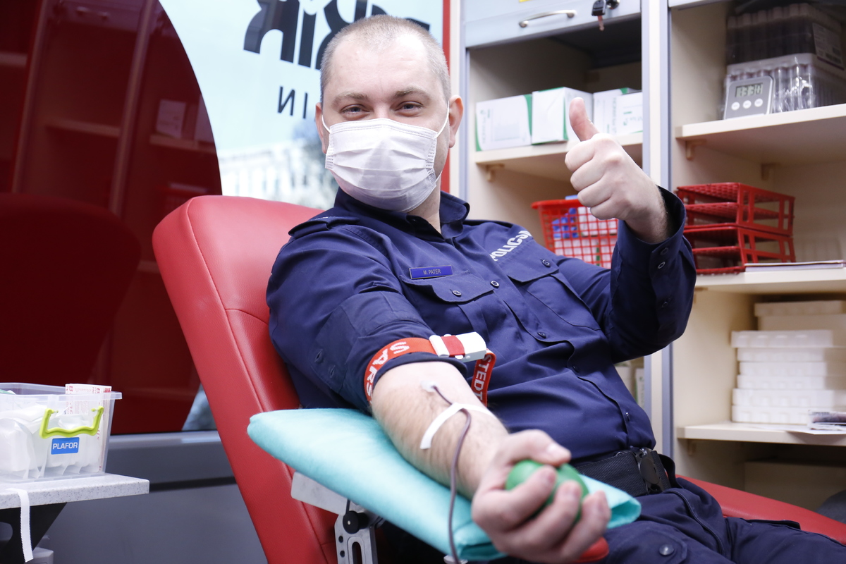 Policjant ubrany w mundur z napisami policja unosi dłoń z wyprostowanym kciukiem w geście zadowolenia w mobilnym punkcie oddawania krwi.