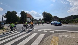 Policjanci w kamizelkach odblaskowych zatrzymali ruch na drodze krajowej numer jedenaście aby rowerzyści mogli bezpiecznie przejechać. W tle widoczny oznakowany radiowóz ustawiony w poprzek drogi.