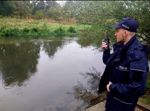 Zdjęcie przedstawia policjanta, rozmawiającego przez radiostację, obok rzeka.