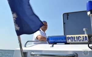 na pierwszym planie flaga ze znakiem policji, w tle policjant na łodzi